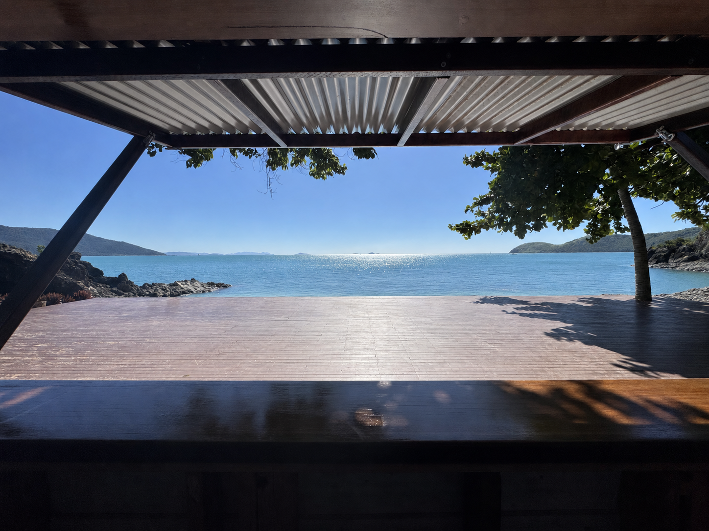 Lovers Cove open-air cocktail bar — timber servery bench and deck framed by a corrugated steel awning, opening out over turquoise Whitsundays water